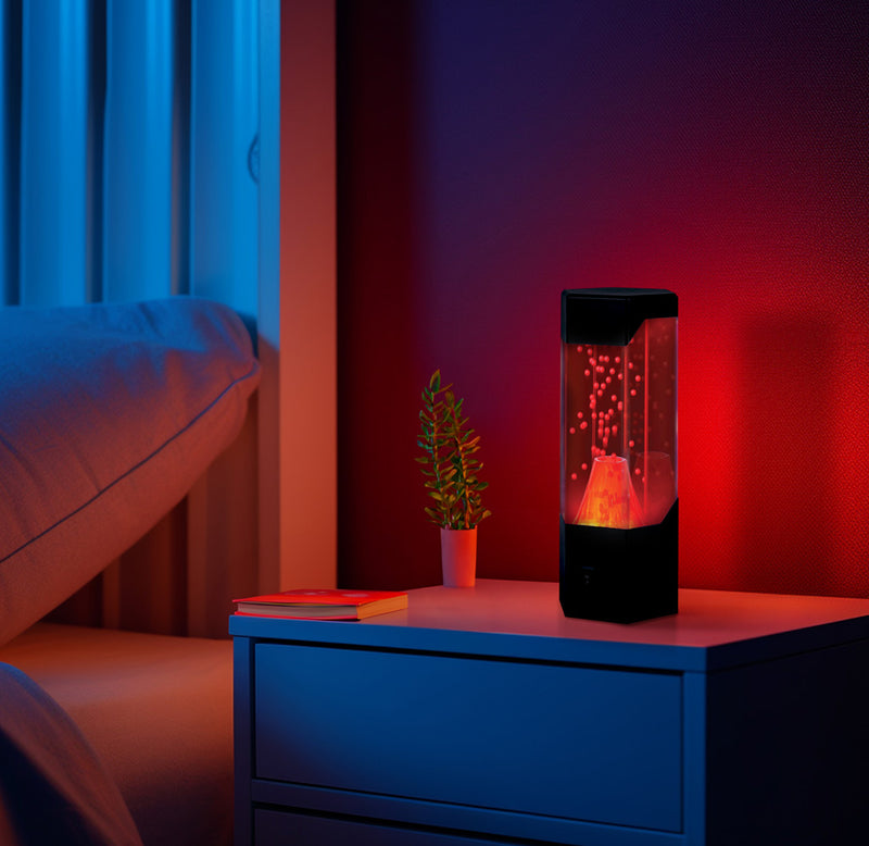 The volcano lamp is turned on and placed on a night stand by a bed in a dimly lit room.