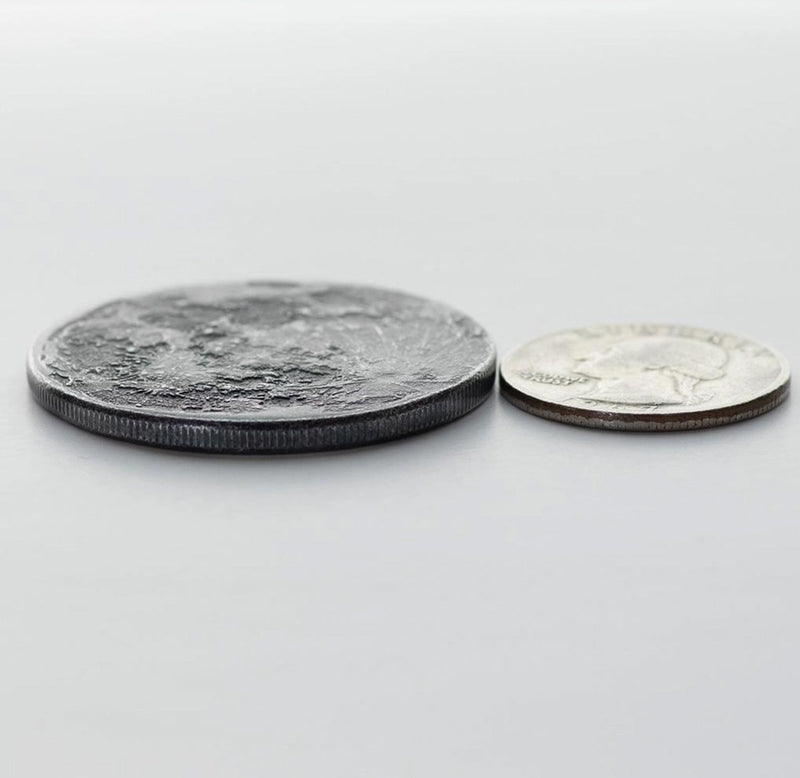 A iron-colored coin that looks like the moon is next to a quarter side by side. 