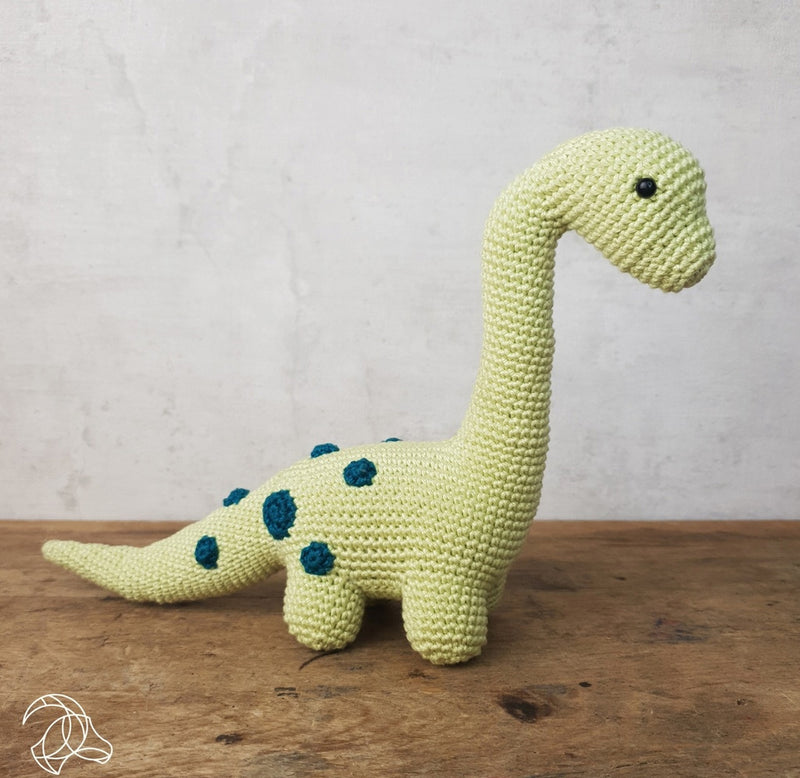 The green crochet brontosaurus is pictured on a wooden table and grey background. 