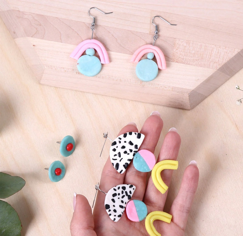 Hands are holding a pair of colorful earrings. There are two other pairs of earrings on a wooden table. 