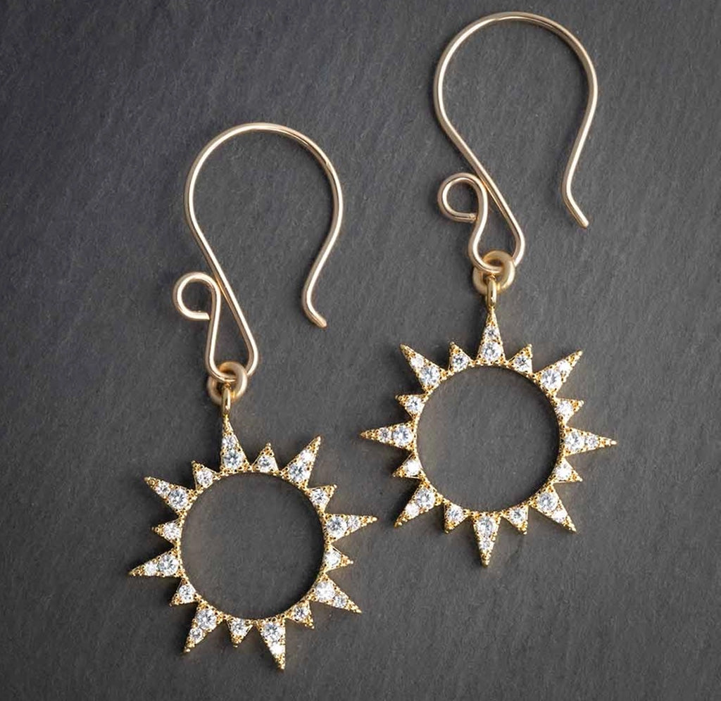 A pair of gold earrings in the shape of a sun with a hollow center. There are white gems lining the sun charm's rays. 