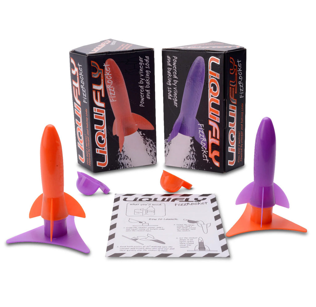 An orange and purple plastic rocket are facing upwards next to a piece of paper. In the back there are two black packages with images of these rockets on them. 