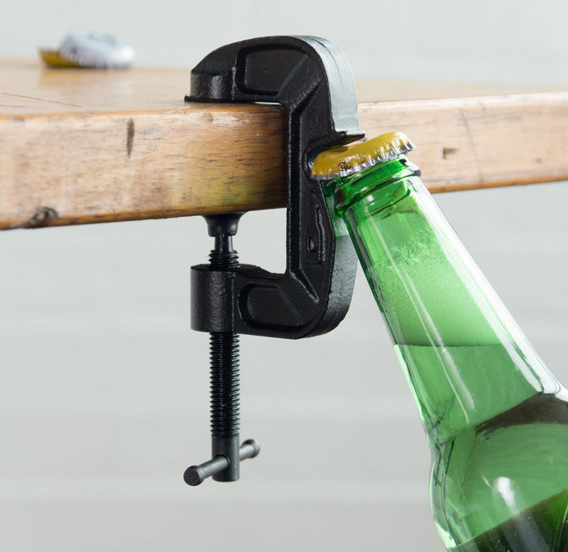 The g-clamp is fastened to the edge of a table and a green bottle is positioned to be opened on the edge of the clamp. 