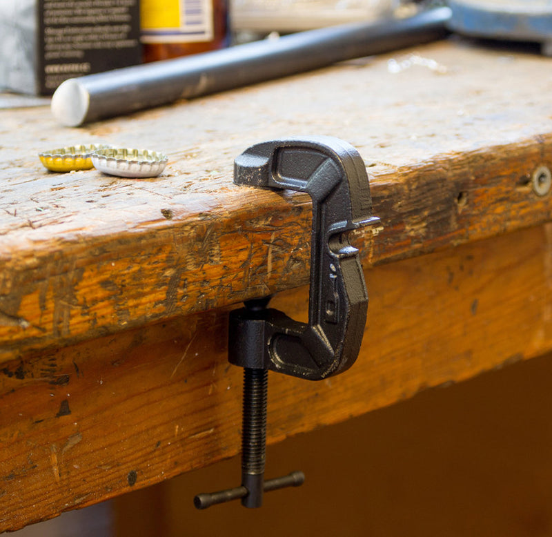 The black g-clamp is fastened to the edge of a wooden table. 