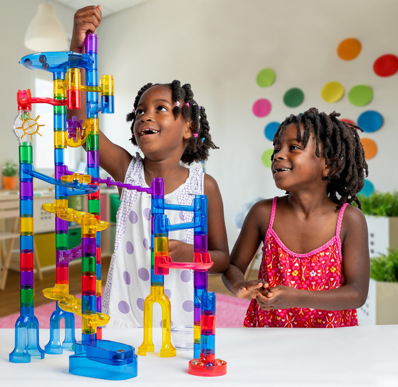 Two children are smiling as they play with a constructed marble run set. One child is adding marbles to the top of the structure while the other is watching the marbles race down. 