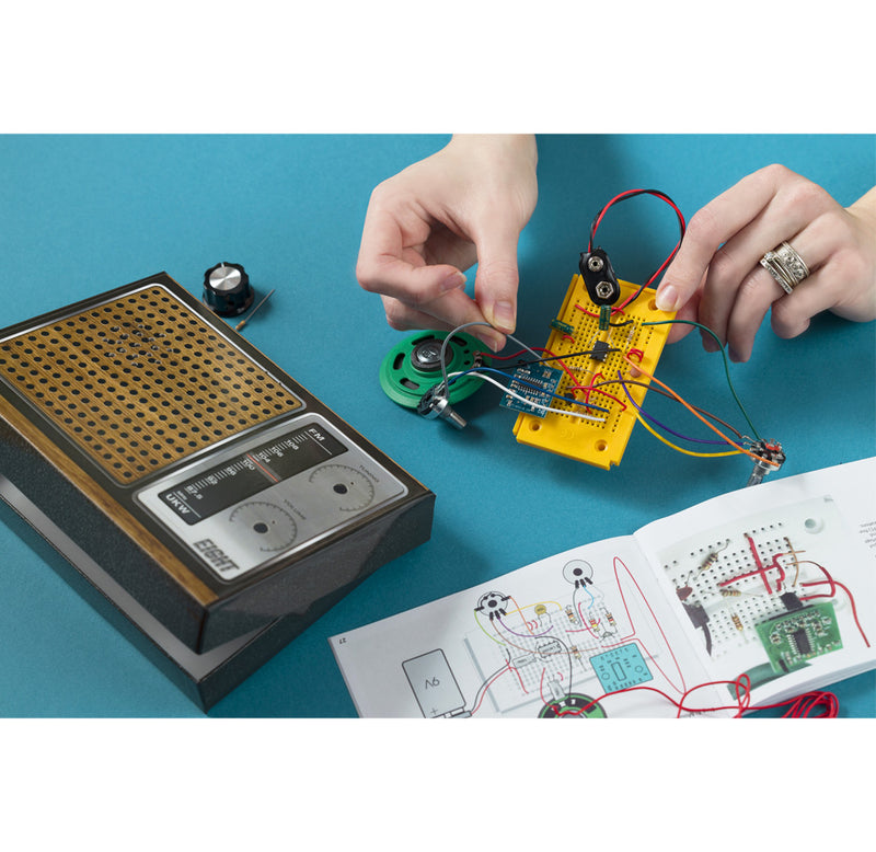 A hand is holding a circuit bard and connecting wires to it. On the table is a box shaped like a radio and an open booklet with instructions. 