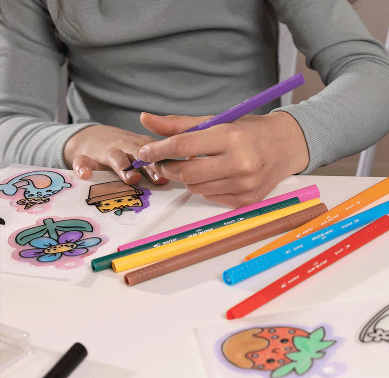 A person is using a purple marker to color in a Shrink-Its illustration. The white table has other illustrations and assorted colored markers. 